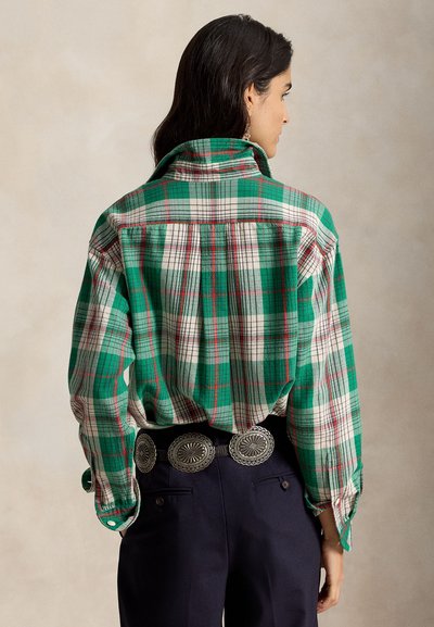 Femme aux cheveux noirs portant une chemise à carreaux verte, blanche et rouge rentrée dans un pantalon bleu marine avec une ceinture noire ornée de grandes conchos argentées.