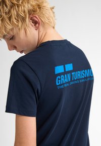 Námorně modré bavlněné tričko s modrým textem "GRAN TURISMO" a "SKUTEČNÝ SIMULÁTOR ŘÍZENÍ" na zadní části, s klasickým kulatým výstřihem.