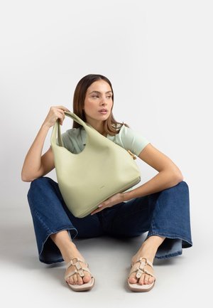 Donna seduta sul pavimento con le gambe divaricate, che tiene una grande borsa verde chiaro, indossa una camicia verde chiaro, jeans blu e sandali beige.