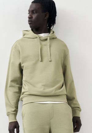 Νεαρός άνδρας με σκουρόχρωμο δέρμα και dreadlocks, που φοράει ανοιχτό πράσινο φούτερ με κουκούλα και αντίστοιχο παντελόνι, κοιτάζοντας αριστερά σε απλό φόντο.