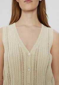 Gebreide vest in lichtbeige met een V-halslijn, voorzien van een knopenfront en een textuurpatroon van strepen en open steken.