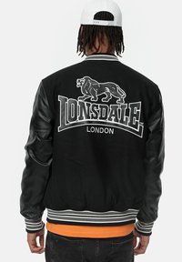 Svart varsityjacka med läderärmar, har en stor lejonlogotyp och texten "LONSDALE LONDON" i vitt, ribbade randiga smockar och nederkant.