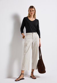 Schwarzes Langarmtop mit V-Ausschnitt, beigefarbene hochgeschnittene Hose, schwarze Riemchen-Sandalen und eine braune Wildleder-Schultertasche.