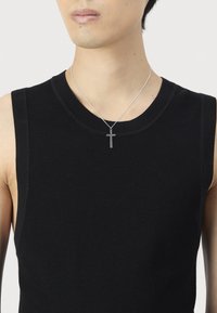 Jaunas žmogus, vilkintis juodą be rankovių megztinį ir sidabro kryžiaus pakabuką, prieš paprastą baltą foną.