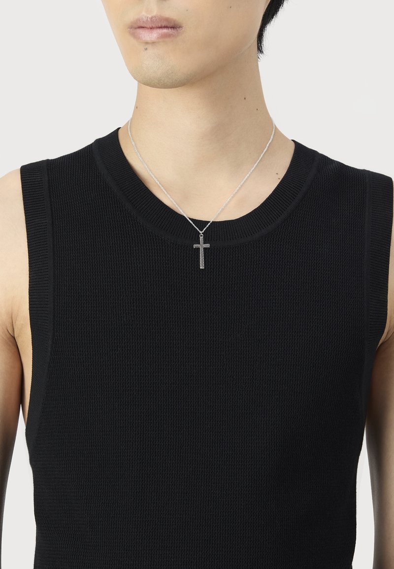 Jaunas žmogus, vilkintis juodą be rankovių megztinį ir sidabro kryžiaus pakabuką, prieš paprastą baltą foną.