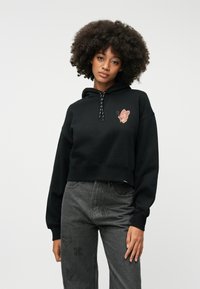 Krótkia czarna bluza z kapturem z grafiką motyla w odcieniach beżu i różu. Posiada sznurki z czarno-białym wzorem, zestawiona z szarymi dżinsami.