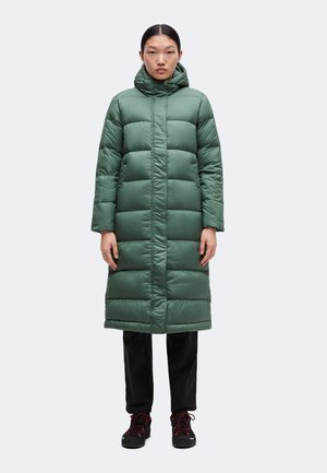 Personne portant un long manteau matelassé vert avec capuche, un pantalon noir et des chaussures de randonnée noires avec des lacets rouges, debout face à l’avant.