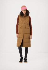 Gilet matelassé marron avec un col haut, fermeture à boutons à l'avant et deux poches. Porté sur un pull couleur rouille et des leggings noirs. Bonnet rose.