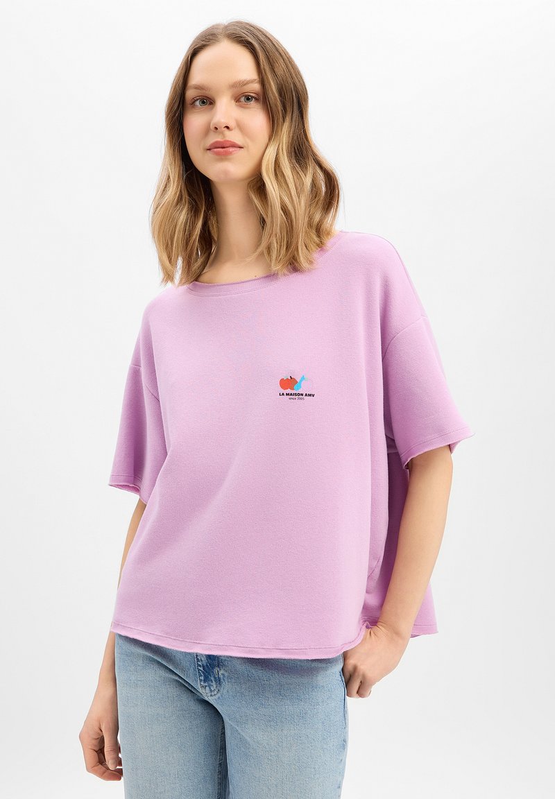 Frau mit schulterlangem welligem Haar, die ein lockeres lavendelfarbenes T-Shirt mit einem kleinen mehrfarbigen Logo und hellblaue Jeans trägt, vor einem weißen Hintergrund stehend.