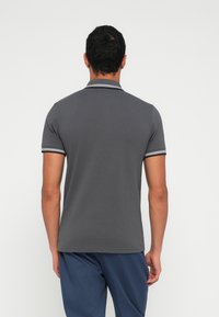 Szara koszulka polo z krótkimi rękawami, kołnierzykiem z czarno-białymi akcentami i gładką fakturą. Noszona z niebieskimi spodniami.