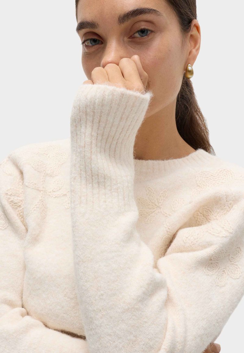 Femme aux cheveux bruns attachés portant un pull en maille crème à motifs floraux, se couvrant la bouche avec la main, une boucle d'oreille en or visible.