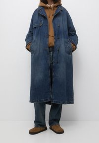 Long manteau en denim avec une finition bleu délavé, cinq boutons et deux poches latérales, porté sur un sweat à capuche marron et un jean. Chaussures marron clair.