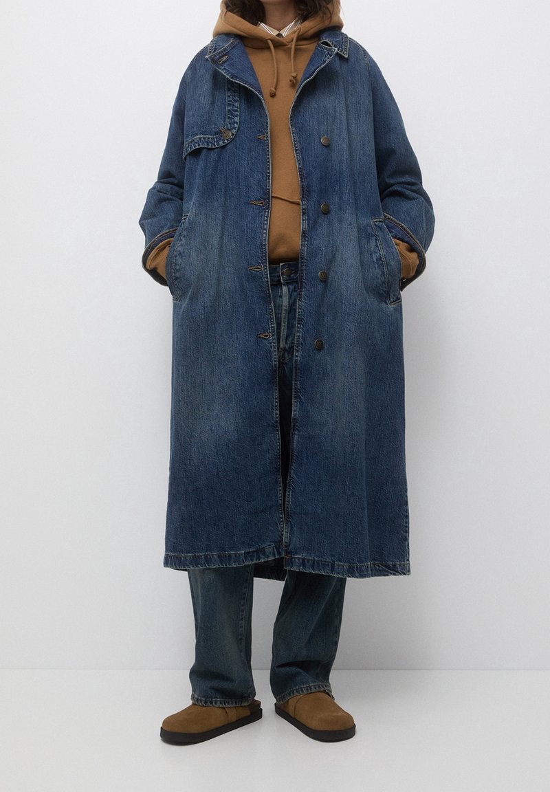 Long manteau en denim avec une finition bleu délavé, cinq boutons et deux poches latérales, porté sur un sweat à capuche marron et un jean. Chaussures marron clair.