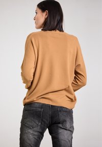 Long-sleeve, camel-colored sweater in soft-knit material, paired with dark gray jeans featuring back pockets and subtle fading.