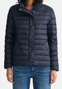 Frau trägt eine marineblaue Steppjacke mit Druckknöpfen und Taschen, kombiniert mit blauen Jeans, steht vor einem einfarbigen Hintergrund.