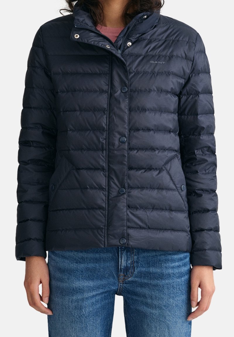 Frau trägt eine marineblaue Steppjacke mit Druckknöpfen und Taschen, kombiniert mit blauen Jeans, steht vor einem einfarbigen Hintergrund.