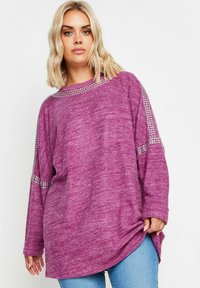 Överdimensionerad lila tröja i en strukturerad tyg med långa ärmar och silverfärgade detaljer längs axlarna. Sömmen är något rundad.