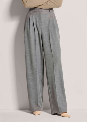 Frau trägt hochtaillierte, weit geschnittene graue Hose und beige spitze Schuhe, steht mit verschränkten Armen vor schlichtem Hintergrund.