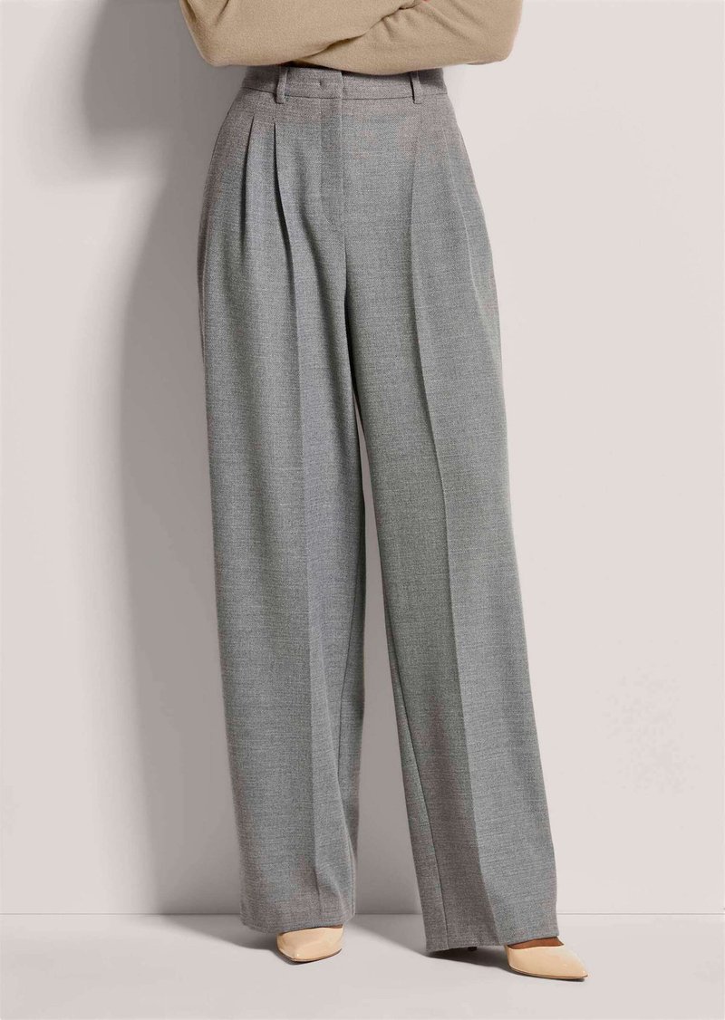 Frau trägt hochtaillierte, weit geschnittene graue Hose und beige spitze Schuhe, steht mit verschränkten Armen vor schlichtem Hintergrund.