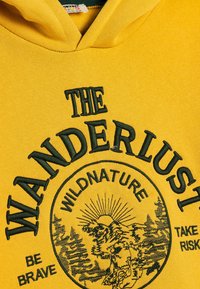 Sudadera amarilla con un gráfico frontal que presenta bordado verde que dice "EL ANHELO DE VIAJAR", con un diseño natural y detalle de estallido solar.