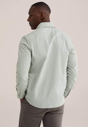 Uomo con capelli corti che indossa una camicia verde chiaro a maniche lunghe e pantaloni grigi, in piedi con le mani nelle tasche, di spalle.