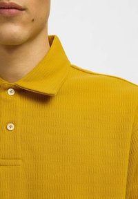 Gelbes Poloshirt mit strukturiertem Muster, geknöpftem Kragen und kleiner Brusttasche. Verfügt über runde, weiße Knöpfe und ribbelige Details.