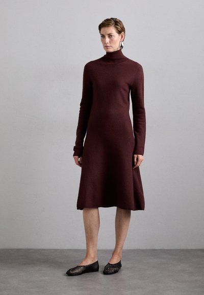 Burgundy turtleneck mekko, jossa on pitkät hihat, kiinnitetty vartalon osa ja levenevä hameosa. Mallilla on päällään mustat verkkokengät, jotka sujahtavat jalkaan. Yksinkertainen, teksturoitu kangas.