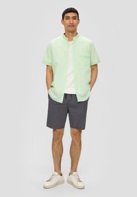 Helles grünes, kurzärmeliges Hemd mit Knöpfen und einer Brusttasche, kombiniert mit dunkelgrauen Shorts und beige Sneakers. Glatte Textur und legeres Design.