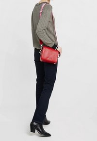 Sac bandoulière rouge en cuir texturé, avec une quincaillerie argentée et une longue bandoulière ajustable. Porté avec un haut à motifs et un jean sombre.