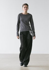 Jeune femme marchant, portant un haut moulant à manches longues gris foncé, un pantalon noir à jambes larges avec une écharpe à motifs, et des chaussures noires.