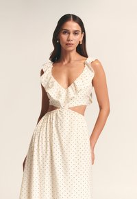 Polka-Dot-Kleid in Creme mit gerafftem Ausschnitt und offenem Mittelteil. Der elastische Taillenbund legt den Stoff, wodurch eine fließende Silhouette entsteht.