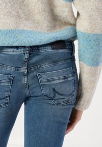 Denim jeans in a medium blue wash with a fitted design, featuring two back pockets and visible stitching details. Soft, textured sweater above.