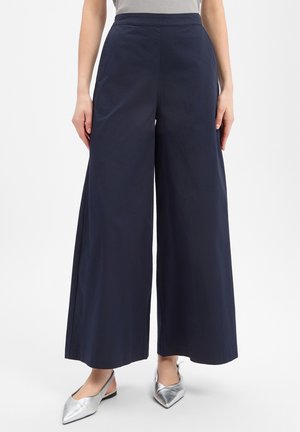 Frau trägt marineblaue weite Hosen und silberne spitze Slingback-Ballerinas, steht vor schlichten Hintergrund.