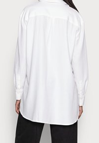 Chemise blanche à manches longues en tissu lisse, avec un empiècement au dos agrémenté d'un pli, des poignets boutonnés et un ourlet arrondi pour une coupe décontractée.