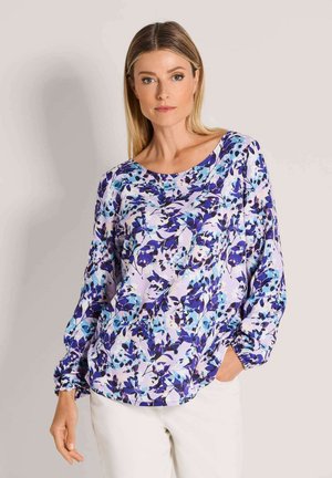 Frau mit glatten blonden Haaren, die eine langärmelige Blumenbluse in violetten und blauen Farbtönen und eine weiße Hose trägt, steht mit einer Hand in der Tasche.