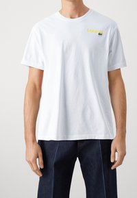 Bílý bavlněný tričko s krátkým rukávem, na levé straně hrudi s žlutým logem "Lacoste" a zeleným výšivkou krokodýla. Klasický střih.