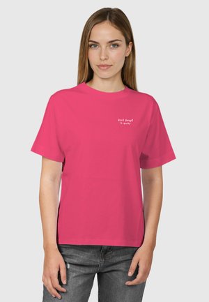 Kobieta o prostych jasnobrązowych włosach, ubrana w jaskraworóżowy T-shirt z napisem „Nie zapomnij się uśmiechnąć” oraz szare dżinsy, na jednolitym tle.