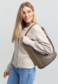 Bruine leren schoudertas met studaccenten, zachte textuur en brede banden, gedragen over een beige cardigan en blauwe jeans.