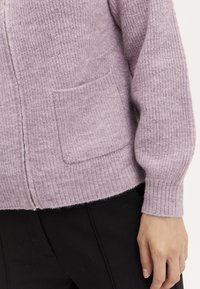 Svetle fialový pletený kardigan s ribbed texturou, má přední kapsu a přiléhavé manžety. Nosený přes černé kalhoty.