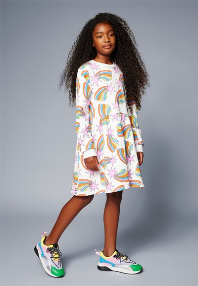 Μακρυμάνικο φόρεμα με πολύχρωμα σχέδια ουράνιου τόξου και αστεριών σε λευκό φόντο, συνδυασμένο με πολύχρωμα πάνινα παπούτσια και μαύρες σόλες.