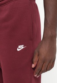 Burgundske Nike trenirka iz mešanice bombaža, ki ima bel logo na levem stegnu in stransko žep z gladko teksturo.