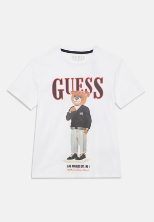 T-shirt blanc à manches courtes avec un motif d'ours portant une veste noire et un pantalon rayé, avec le texte "GUESS" en rouge au-dessus.