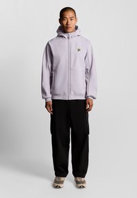 Lyslilla zip-up jakke med hætte, der har et lille gult logo. Bæres sammen med sorte cargo-bukser og beige sneakers. Glat tekstur.