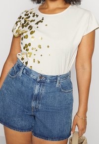 Kvinna iklädd vit T-shirt med guldiga abstrakta prickar och högmidjade blå denimshorts, som håller en beige vävd handväska i höger hand.