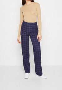 Kvinna som bär en beige långärmad topp, marinblå raka byxor med ett ljust rutmönster och vita sneakers, stående mot en enfärgad bakgrund.