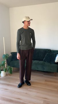 Long-sleeve olive green shirt with a small logo, brown wide-leg trousers, and black shoes, standing on light wooden floor by a green sofa.