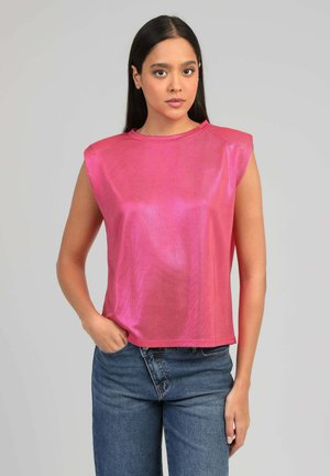 Jeune femme aux cheveux longs et noirs portant un haut sans manches rose vif et un jean bleu, debout avec une main dans la poche.