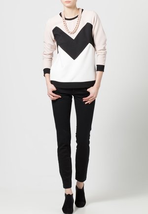 Person standing wearing a pale pink, black, and white sweater with a large chevron design, black pants, black ankle boots, and a chunky necklace.