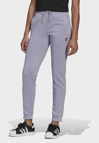 Osoba v světle fialových teplácích Adidas se stahovacími šňůrkami a černých teniskách Adidas s bílými pruhy na jednobarevném pozadí.