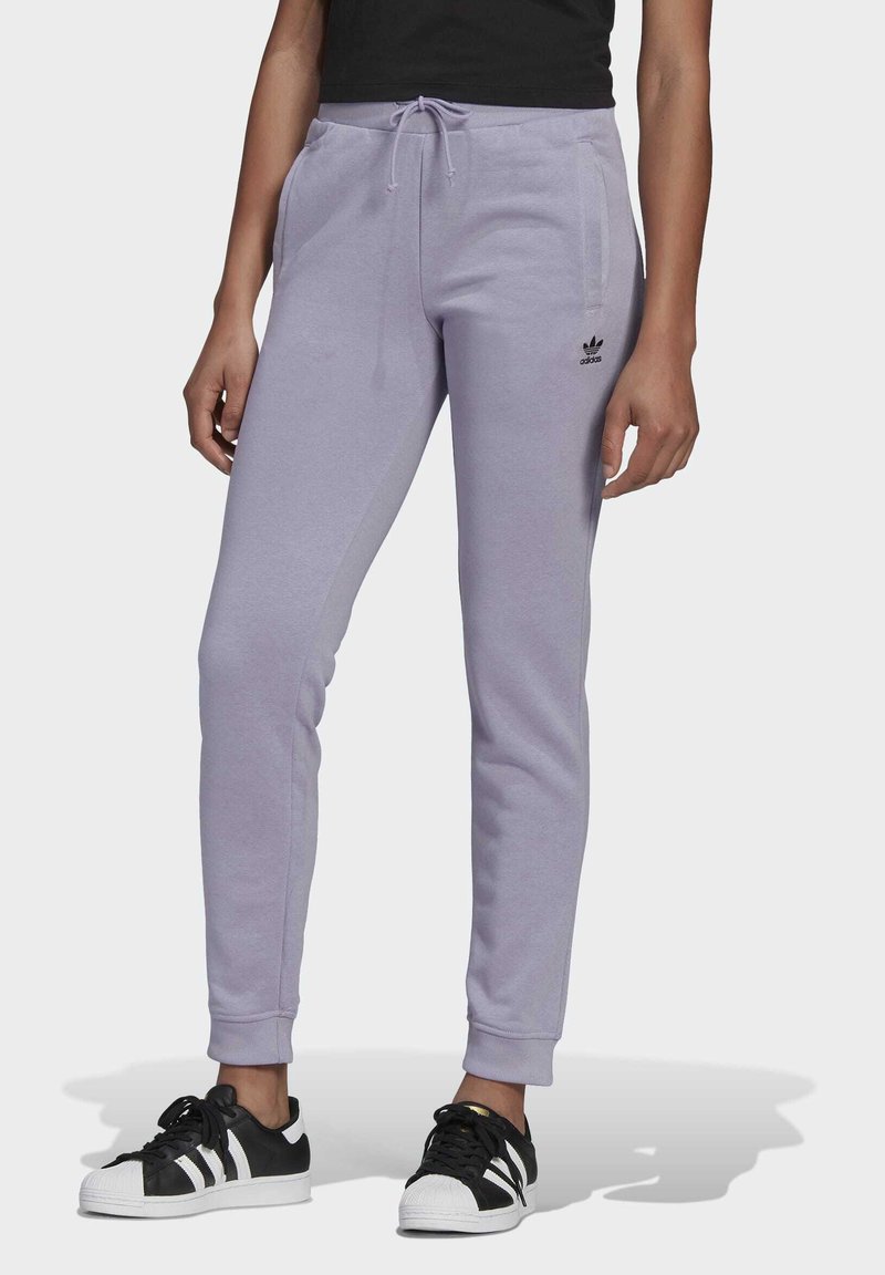 Osoba v světle fialových teplácích Adidas se stahovacími šňůrkami a černých teniskách Adidas s bílými pruhy na jednobarevném pozadí.